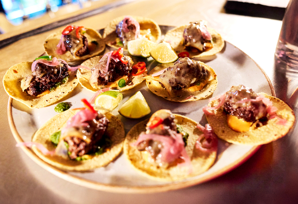 A plate of small tacos topped with meat, pickled onions, sliced chilies, and sauce, garnished with lime wedges, offers a tasty break for those navigating London student living costs—perfect for a student living guide on affordable eats.