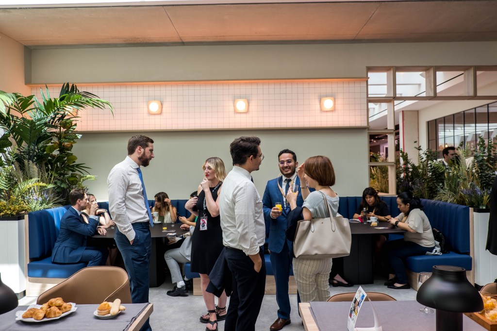 A group of people in business attire are standing and talking in a modern, bright café, while others sit at blue booths eating and conversing. The lively atmosphere encourages desarrollo personal and fortalece habilidades blandas among guests.