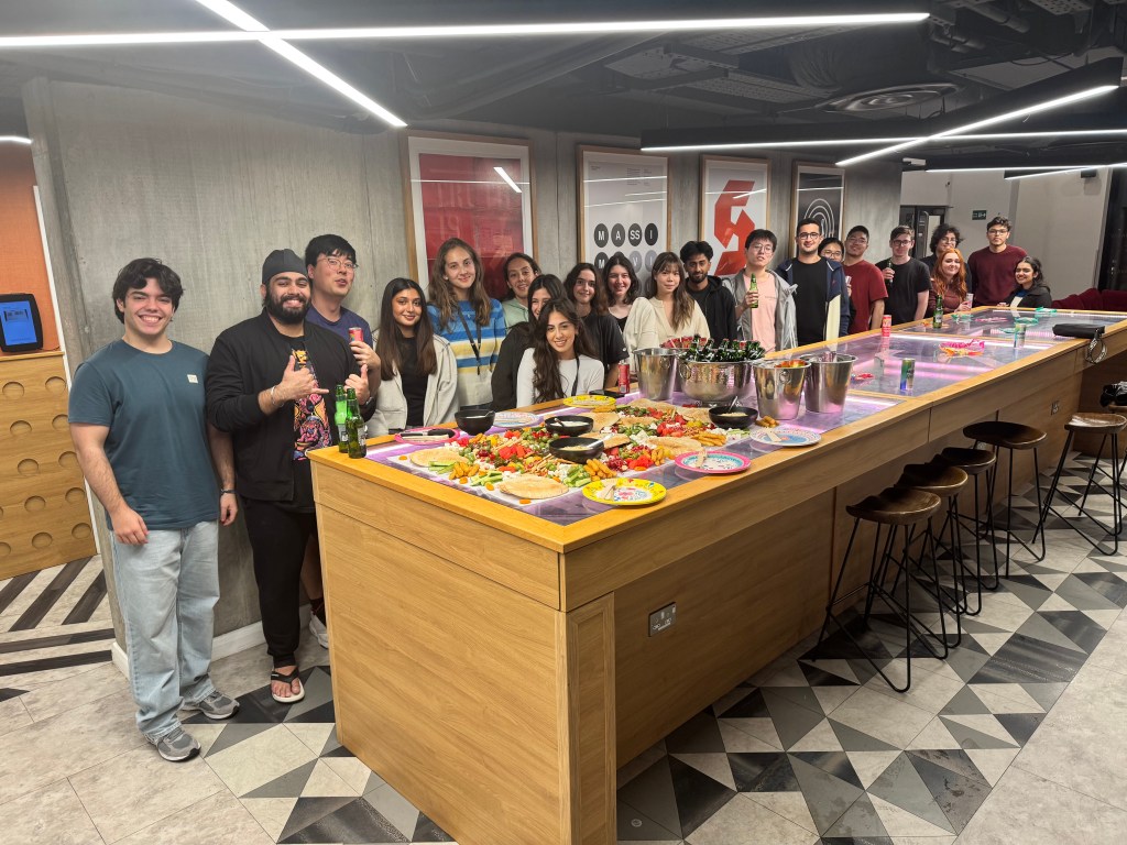 A group of people smiles and poses around a long table filled with colorful food and drinks in a modern indoor setting, celebrating su desarrollo personal en un ambiente vibrante y acogedor.