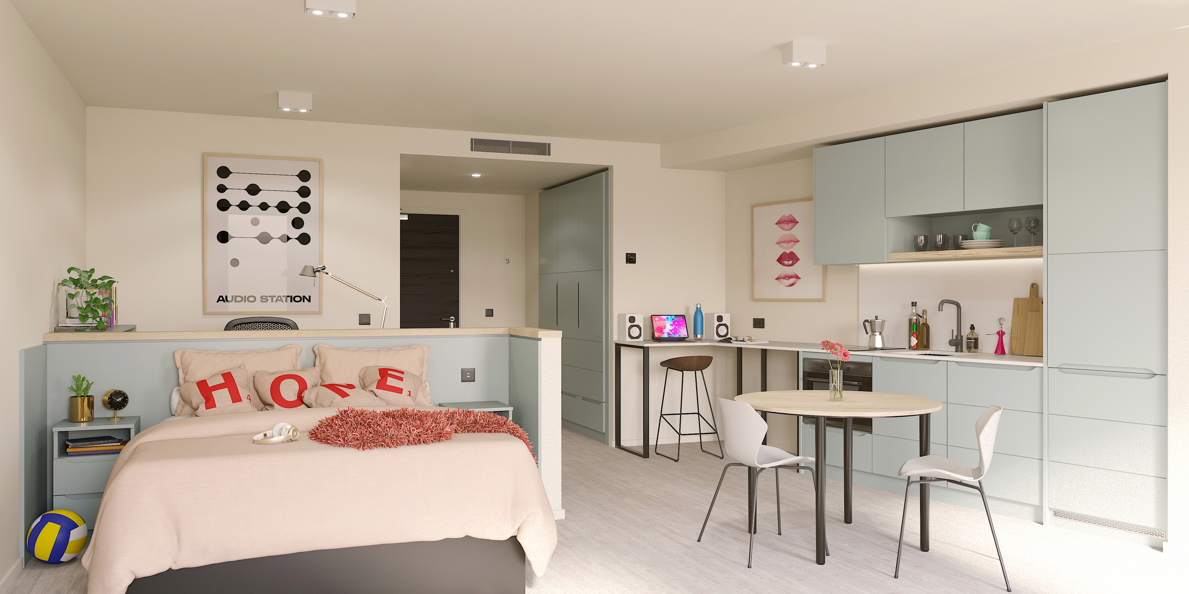 A modern studio apartment with a bed, pastel blue kitchen, small dining table, and cozy decor. The bed has pillows spelling "HOME," art on the wall, and the kitchen area includes cabinets, a sink, and a breakfast bar.