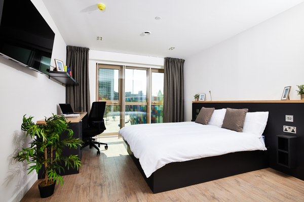 A modern, bright Lewisham bedroom with a large bed, desk and chair—perfect for the best study spots—plus a flat-screen TV, potted plant, and floor-to-ceiling windows leading to a balcony overlooking city buildings.