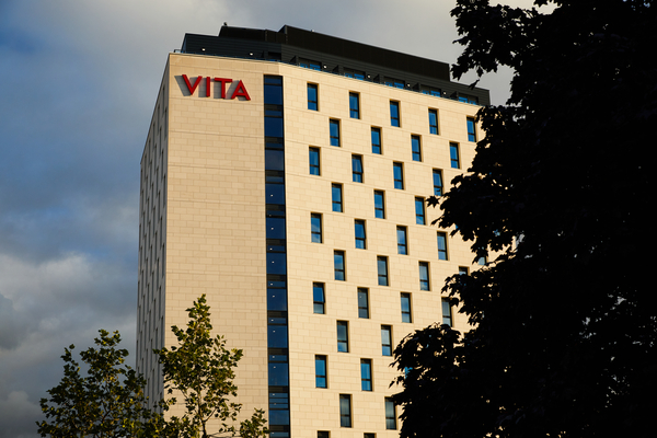 A tall modern building with cream-colored walls and many windows, featuring the red "VITA" sign near the top. Perfect for living close to campus in Leeds, with trees partially obscuring the lower right corner under a cloudy sky.
