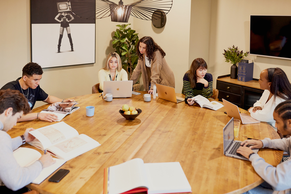 Six young adults gather around a large wooden table in one of the best study spots Lewisham has to offer, working on laptops and reading books. The modern room features art, plants, and a bowl of fruit for a comfortable study vibe.
