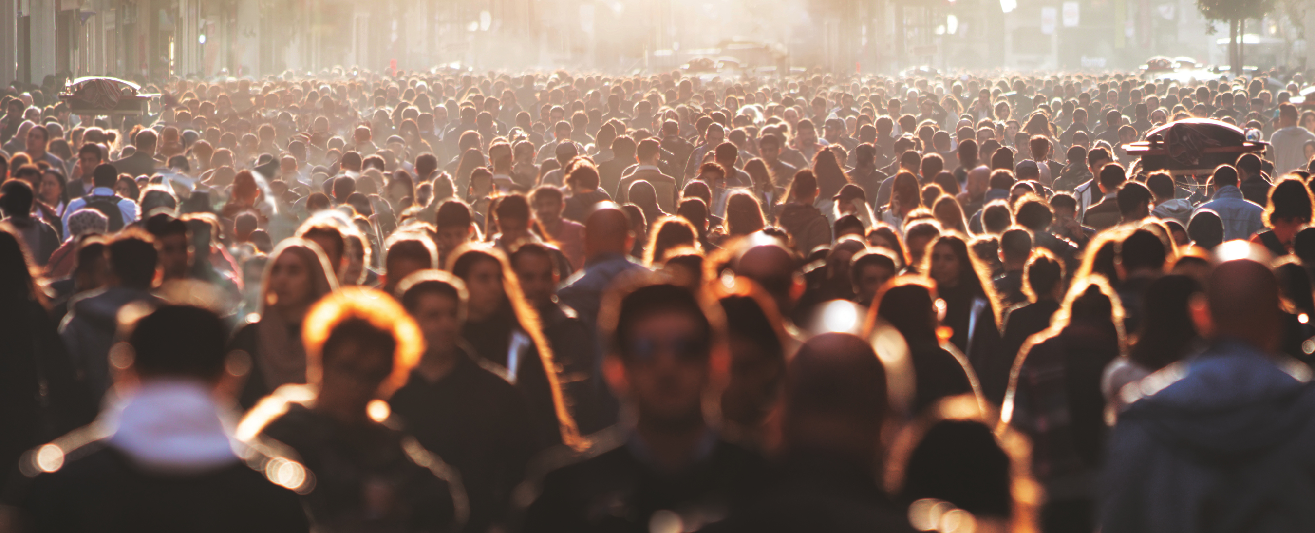 A large crowd of people walks along a city street in the early morning or late afternoon, with sunlight creating a warm, glowing effect and casting long shadows. The vibrant scene hints at the city's bustle and blurred faces moving toward vital mental health services.