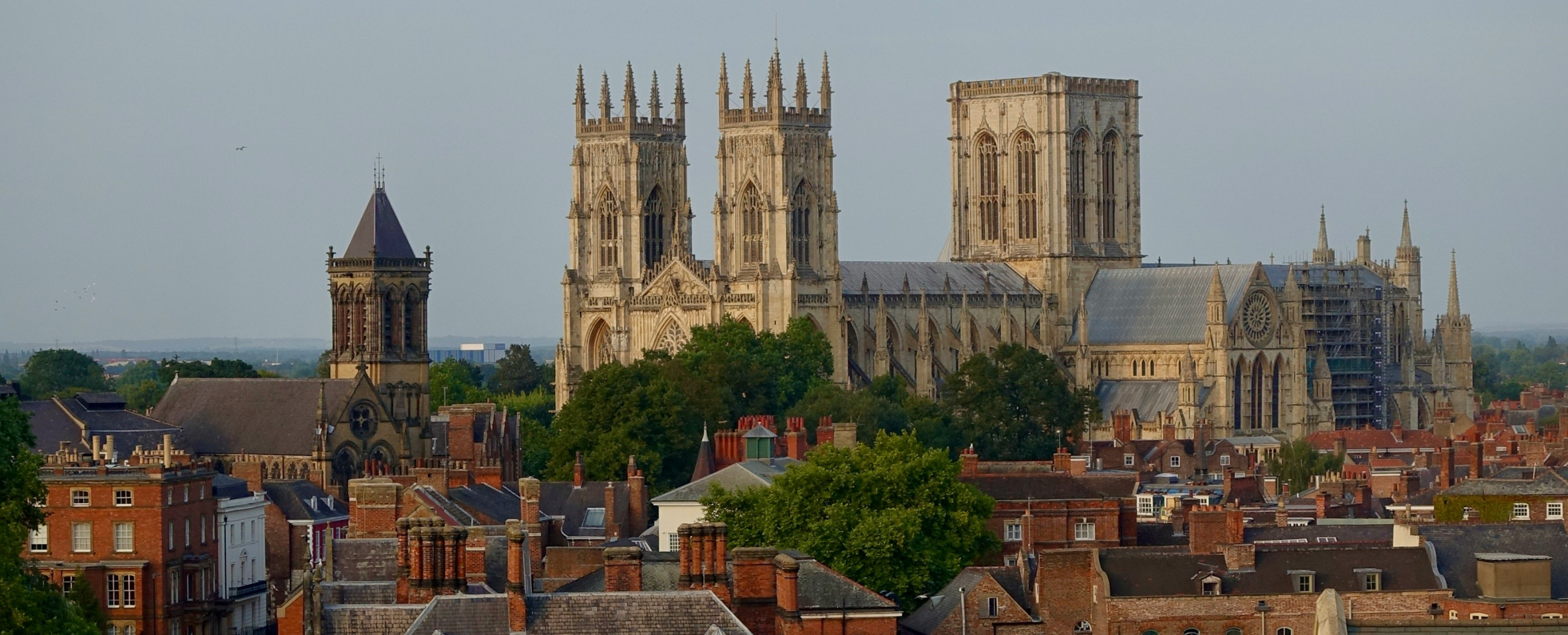 A panoramic view of York Minster, a large Gothic cathedral, rising above red-roofed buildings and trees in York, England—an inspiring sight near some of the best study spots York has to offer for University of York students, under a clear sky.