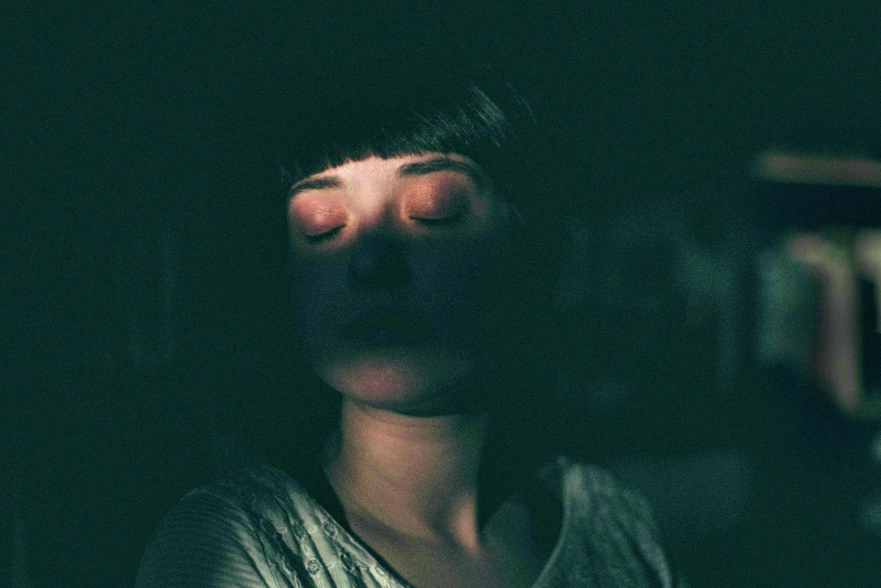 A person with short dark hair stands in partial darkness, eyes closed, with a sliver of light illuminating their face and upper chest—a calm and introspective moment reflecting on salud mental, much like estudiantes at Vita Student Barcelona.