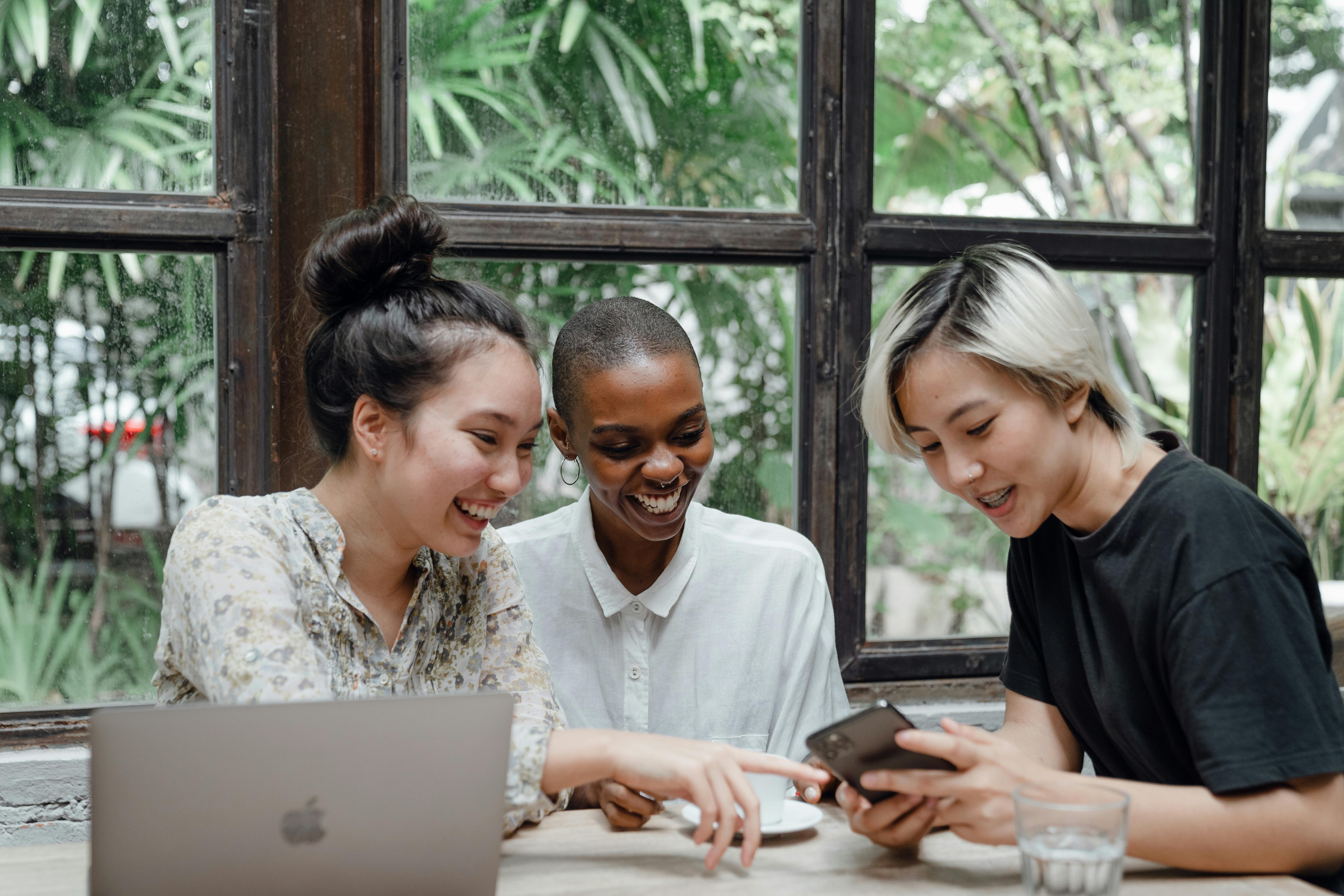 Three people sit at a wooden table near a window, smiling and looking at a smartphone. A laptop and glass of water are on the table, while outside, greenery sets the scene—they could be discussing desarrollo personal or estudiar en el extranjero.
