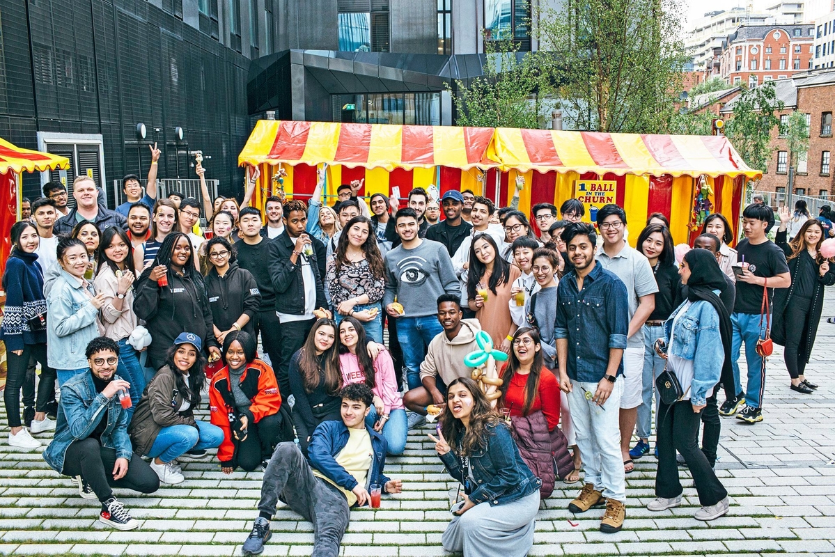 A large, diverse group of young adults, future Vita Student alumni, poses and smiles outdoors in front of a red and yellow carnival tent. Some hold drinks or balloons, celebrating lifelong friends with city buildings in the background.