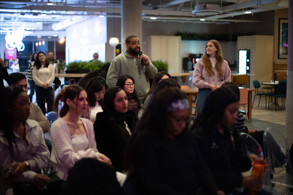 A man stands and speaks into a microphone in a brightly lit room, while a woman nearby smiles. Several people sit in rows, attentively listening—perhaps learning how to create an ATS-friendly CV for UK employers in this modern office setting.