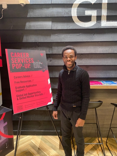 A man in a black sweater and pants stands smiling next to a red and pink “Career Services Pop-Up” sign in a modern, brightly lit space—offering tips on ATS-Friendly CV optimization for UK employers.