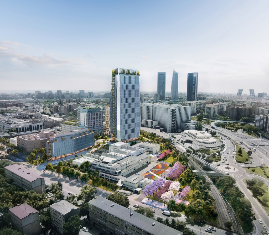 Aerial view of a modern cityscape featuring the tall white Vita Student Oria skyscraper with a rooftop garden, surrounded by contemporary buildings, tree-lined streets, and landscaped parks, with more high-rises visible in the distance.
