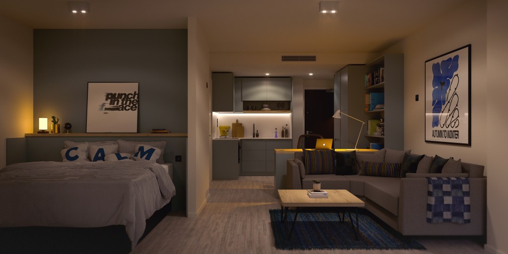 Modern studio apartment at Vita Student Oria with a cozy bed on the left, a gray sofa and coffee table in the center, and a small kitchen with bright lighting in the background. Artworks and decorative pillows add a stylish touch.