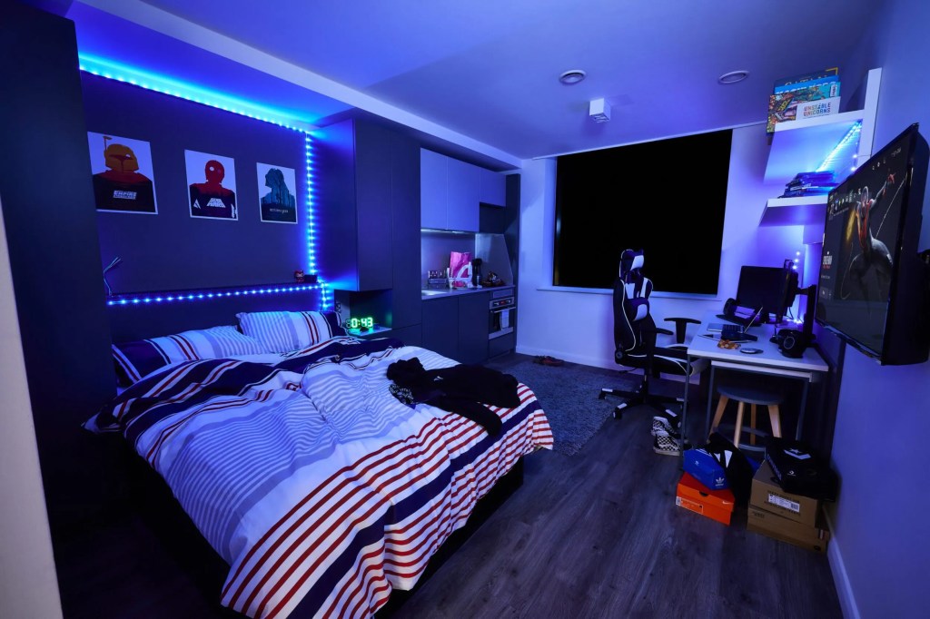 A modern student room featuring blue LED lights, a neatly made bed with striped bedding, wall-mounted TV, gaming chair, desk with computer setup, and superhero posters. The uncovered window reveals nighttime outside—student decor that makes it feel like home.