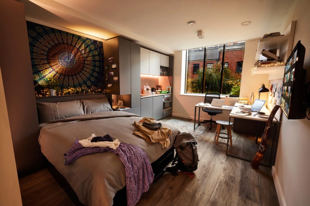 A cozy, modern student room with a large bed, clothes on the bed, a tapestry above the headboard, and a spacious window. With student room decor and warm lighting, it’s designed to feel like home—complete with a desk, laptops, and a guitar by the wall.