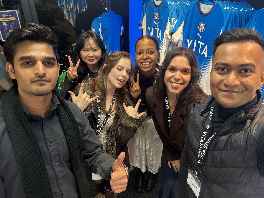 Six people smiling and posing for a selfie in a sports merchandise shop, with blue and white soccer jerseys in the background. Wearing event lanyards, they capture a moment as lifelong friends for the Alumni Archives.