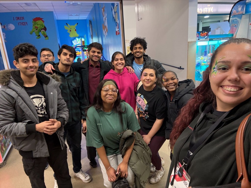 A group of ten smiling young adults, future Vita Student alumni, pose together indoors at an arcade. Colorful wall art and arcade machines fill the background as one person with face paint holds the camera for a selfie—moments with lifelong friends.