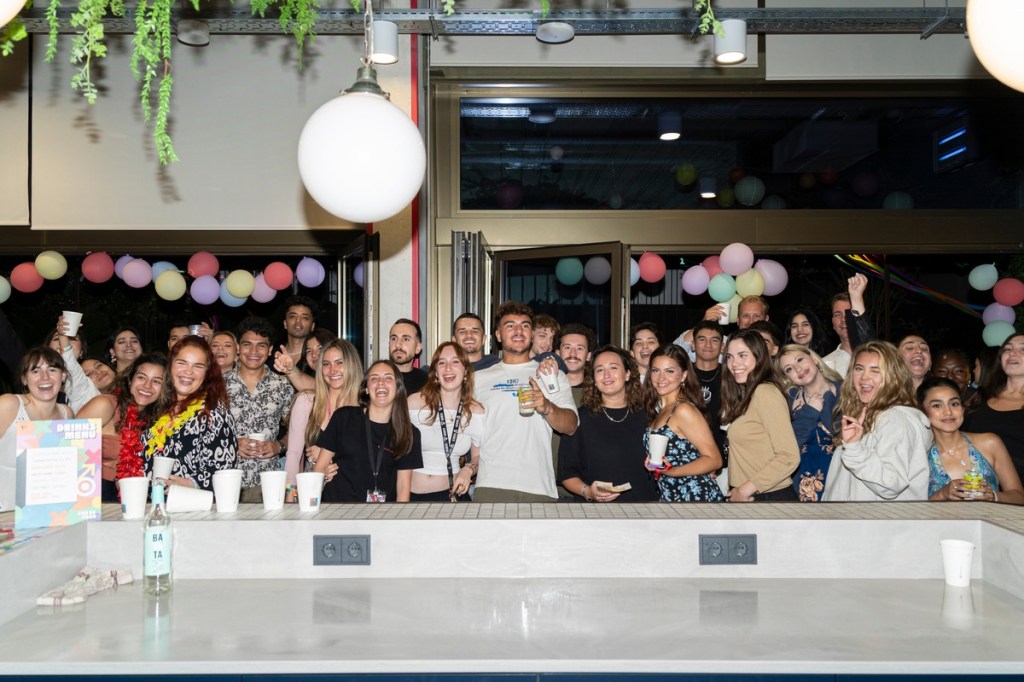 A large group of people stand smiling and posing for a photo at a lively Vita Student indoor party with colorful balloons and decorations. Some hold drinks while others wave or gesture happily, capturing memories as lifelong friends.