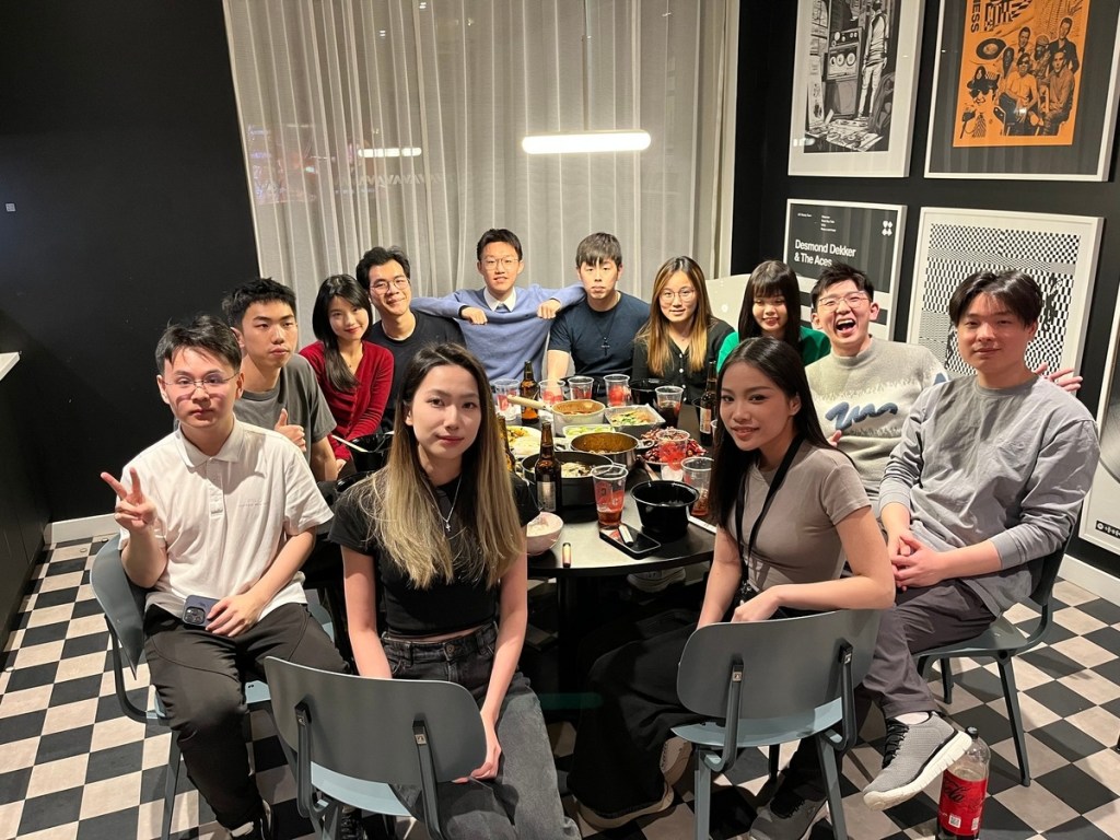 A group of twelve young adults, possibly lifelong friends, are seated around a dining table filled with food in a modern restaurant with checkered flooring and framed art, smiling and posing for the Vita Student Alumni Archives.