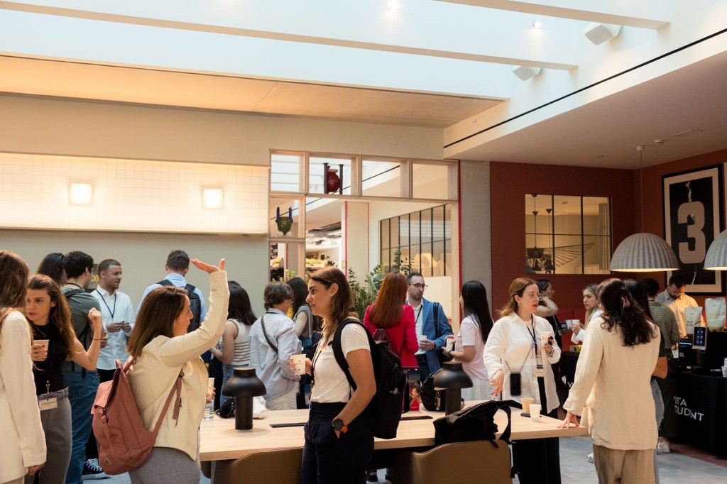 A group of people socialize in a modern, well-lit Vita Student space with large windows. Some are standing and chatting, while others hold drinks. The atmosphere is casual and lively, perfect for making lifelong friends amid contemporary decor.