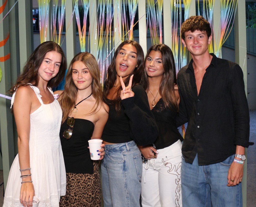 Five young people pose together and smile in front of colorful streamers; four women stand on the left, one holding a cup and another making a peace sign, and a man stands on the right—all celebrating like amigos para toda la vida in casual summer outfits.