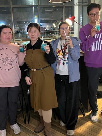 Four people stand indoors at Vita Student, smiling and holding blue drinks with straws. Casually dressed, with two wearing glasses, they enjoy vibrant student life amid large windows and modern hanging lights.