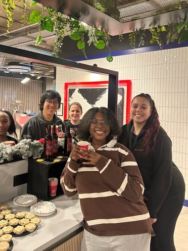 Five people smiling and posing together indoors by a festive counter with drinks and a tray of small pies, enjoying December events. Plants hang above and a holiday decoration lines the counter, capturing the spirit of student life.