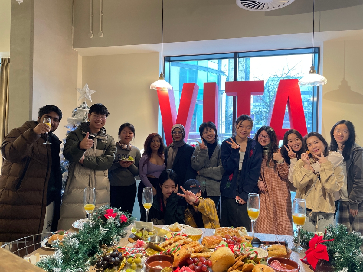 A group of people smiling and posing together indoors in front of a festive food table at a December at Vita Student event. Behind them is a large red "VITA" sign and a decorated Christmas tree by the window.