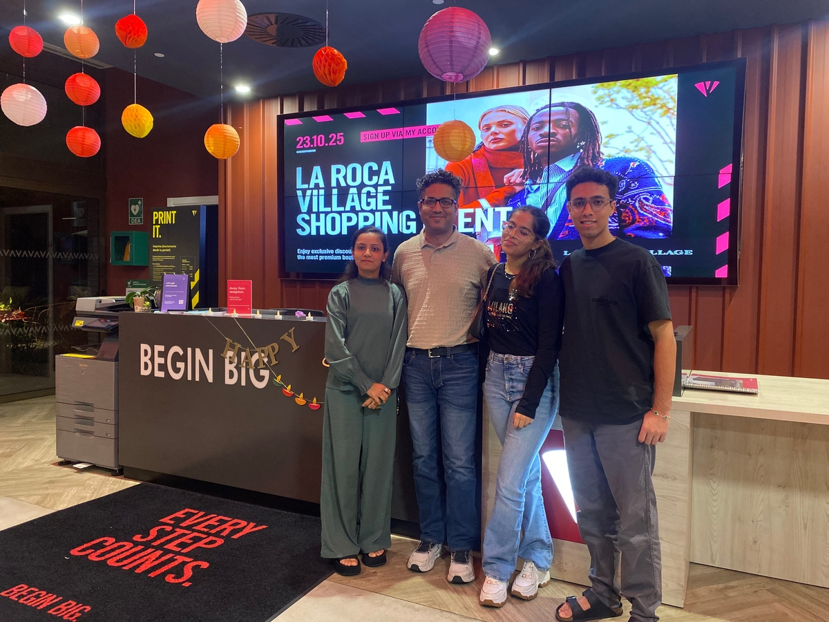 Four people stand and smile in front of a "BEGIN BIG" reception desk at Vita Student. Behind them, a digital screen promotes the La Roca Village Shopping Event, while colorful lanterns set the mood for the upcoming Social Media Career Workshop.