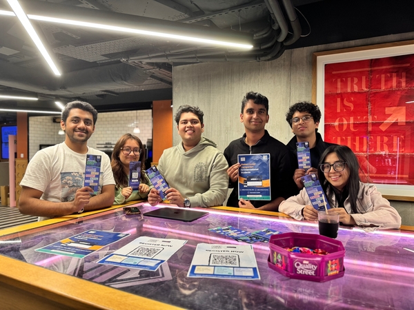 Six young adults stand smiling behind a table at Vita Student, holding up flyers and bookmarks. On the table are printed materials about a social media career, a QR code, and Quality Street chocolates. A red poster hangs on the wall behind them.