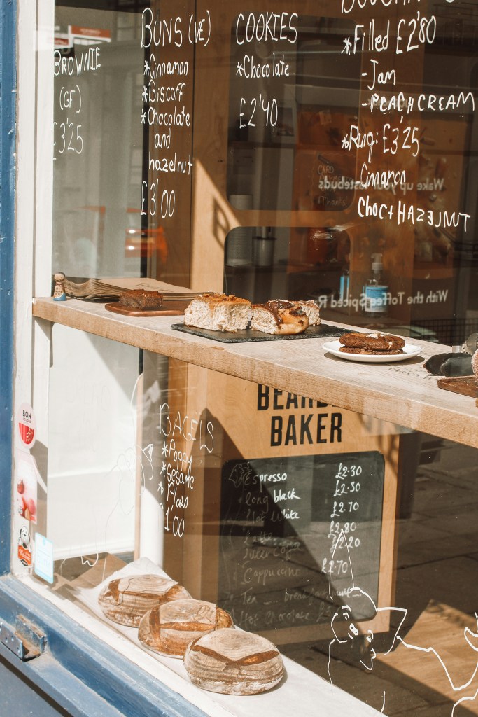 A bakery window display in Edinburgh with various breads, pastries, and cookies on wooden shelves. Menu items and prices are handwritten on the glass in white, listing brownies, buns, cookies, and bagels—perfect for a study break.