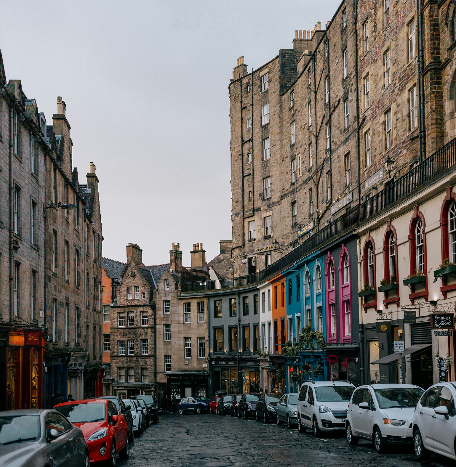 A cobblestone street curves uphill between historic stone buildings in Edinburgh, with parked cars lining both sides. Some shopfronts are painted in bright colors, and the sky is overcast—a scene perfect for those who wish to study Edinburgh’s charm.
