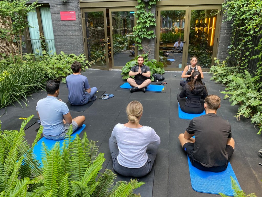 A group of people sit on blue yoga mats in an outdoor garden, which feels like a serene student room, facing two instructors who are leading a meditation or yoga session amid lush green plants and vines, with glass doors in the background.