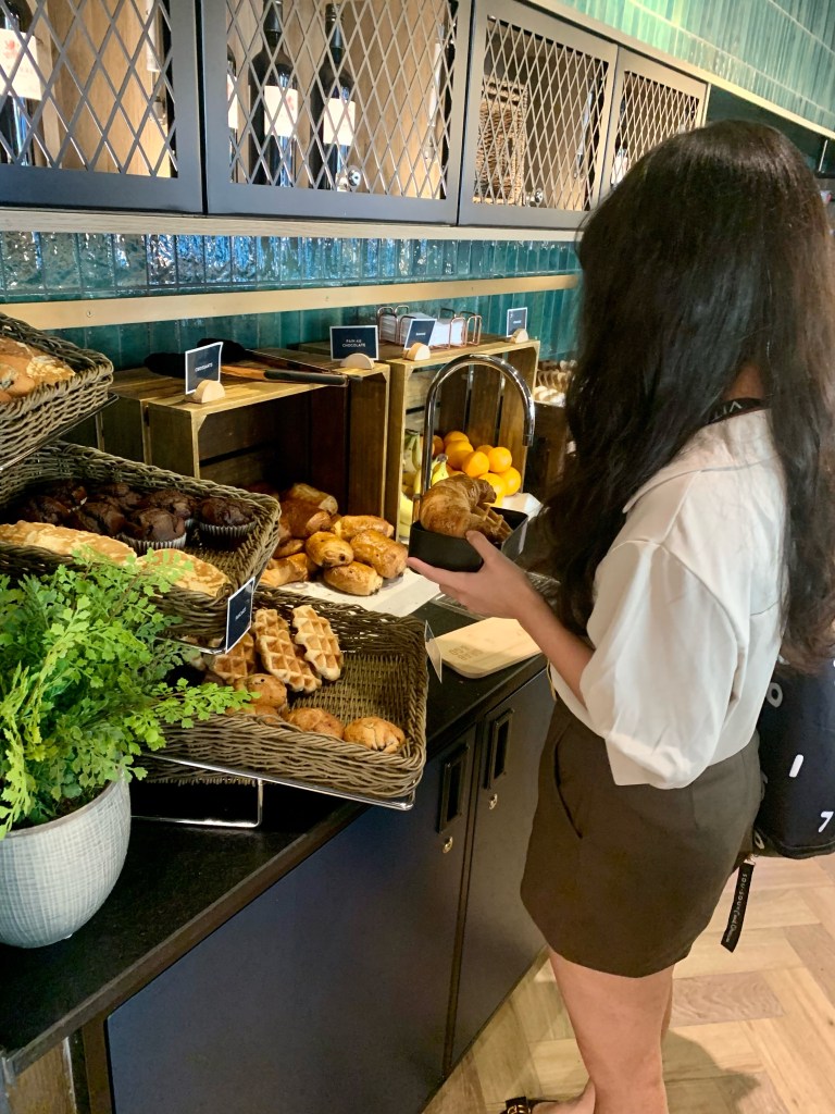 A woman with long dark hair selects a pastry from a buffet table filled with assorted baked goods in a cozy student room, decorated with a potted plant and containers of oranges nearby.