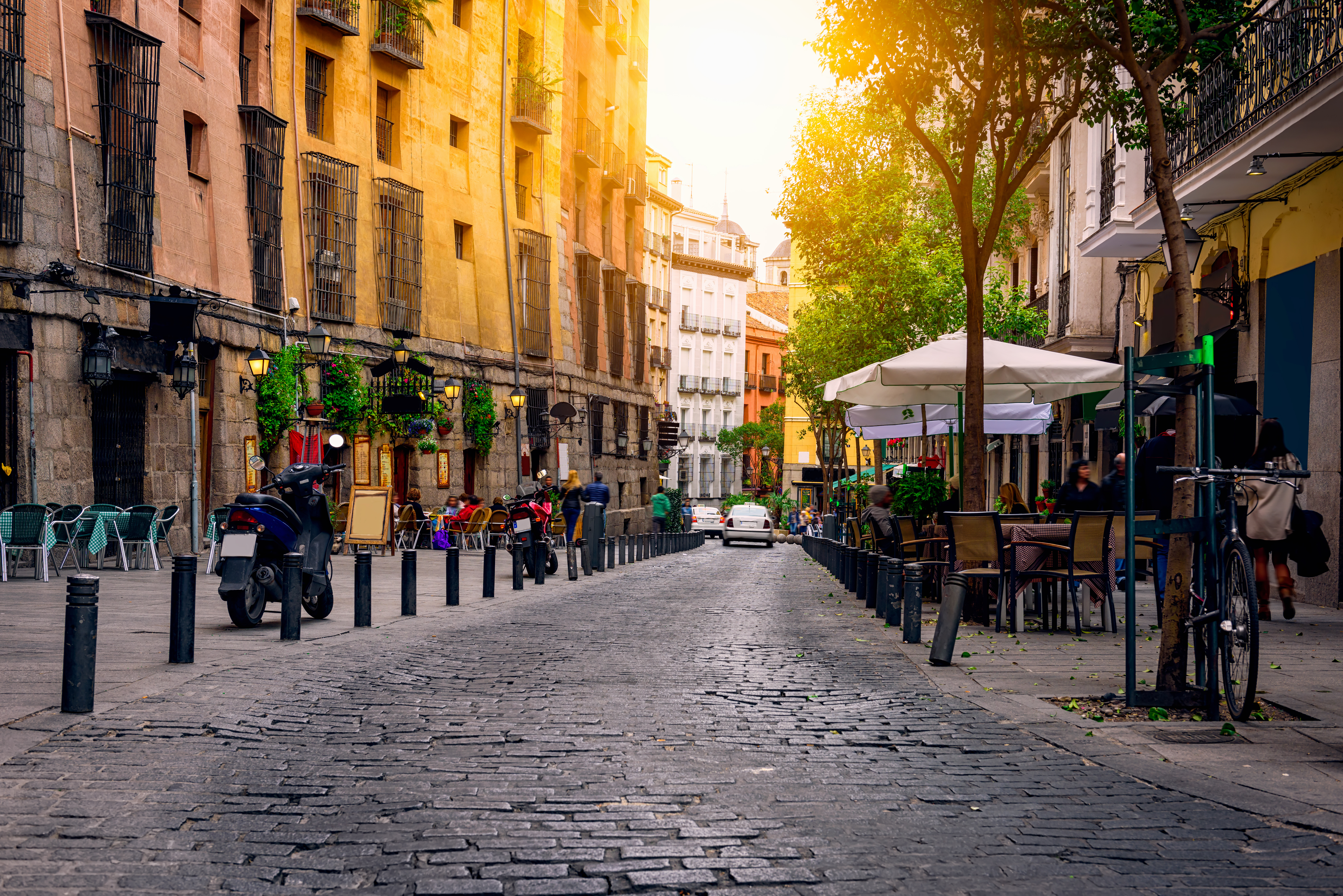 A cobblestone street in Madrid lined with colorful buildings, outdoor cafes, and parked scooters—one of the best places to study for IE Business School students, with people enjoying tables under umbrellas in the warm, golden sunlight.