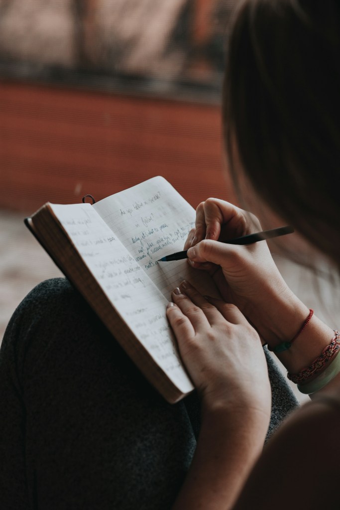 A person sits in a student room with a notebook on their lap, writing with a pen. The journal pages are filled with handwritten text, and the scene feels calm and introspective.