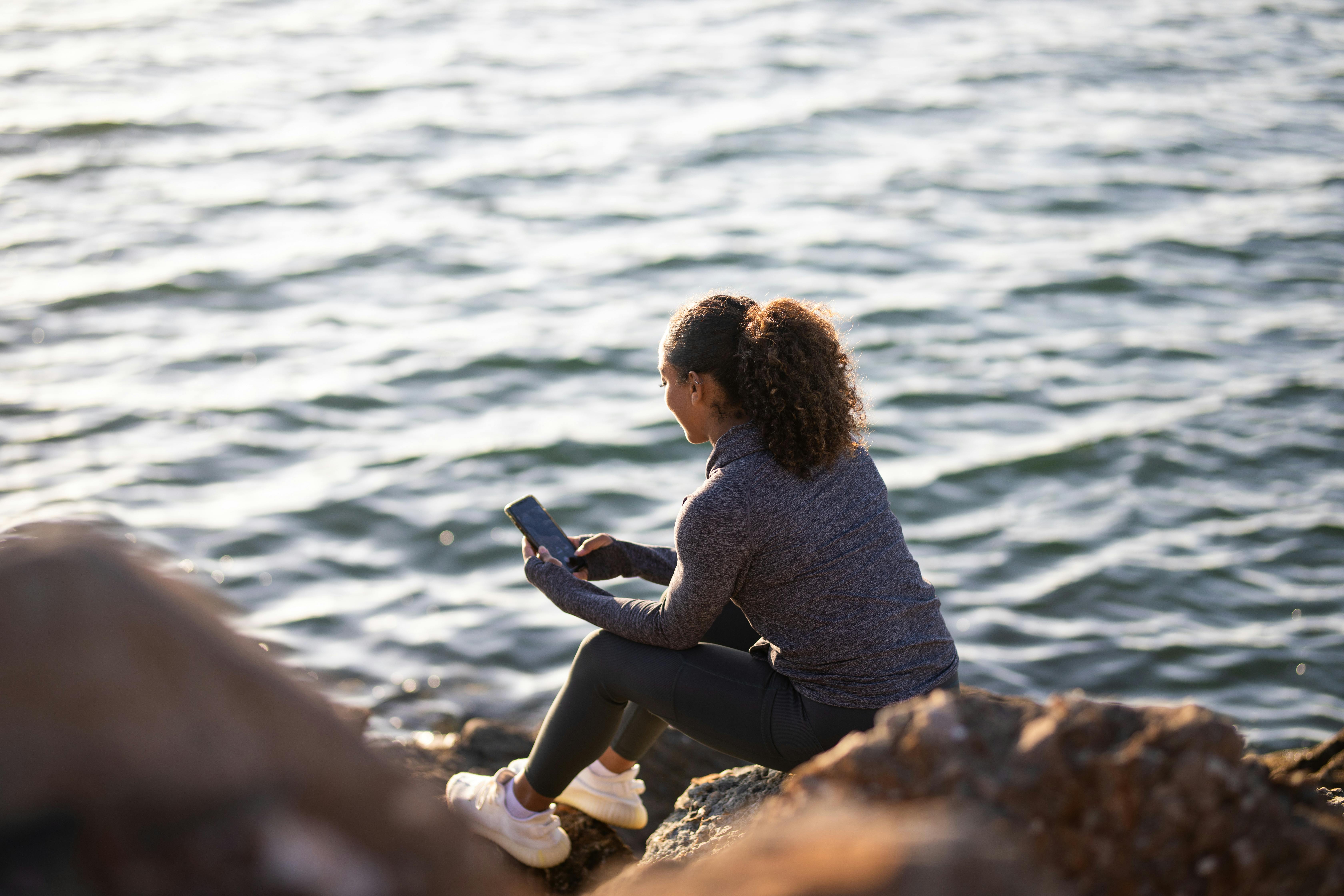 A woman sits on rocks by the water, dressed in athletic clothing and sneakers, smiling at her phone—perhaps checking messages from her student room—as sunlight reflects off the rippling surface behind her.