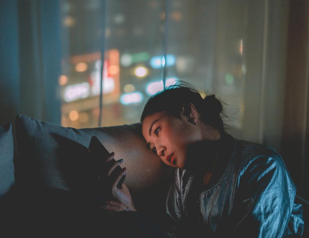 A young woman sits on a couch at night in her student room, looking thoughtfully at her phone. City lights are blurred in the background through a window, creating a moody, urban atmosphere.