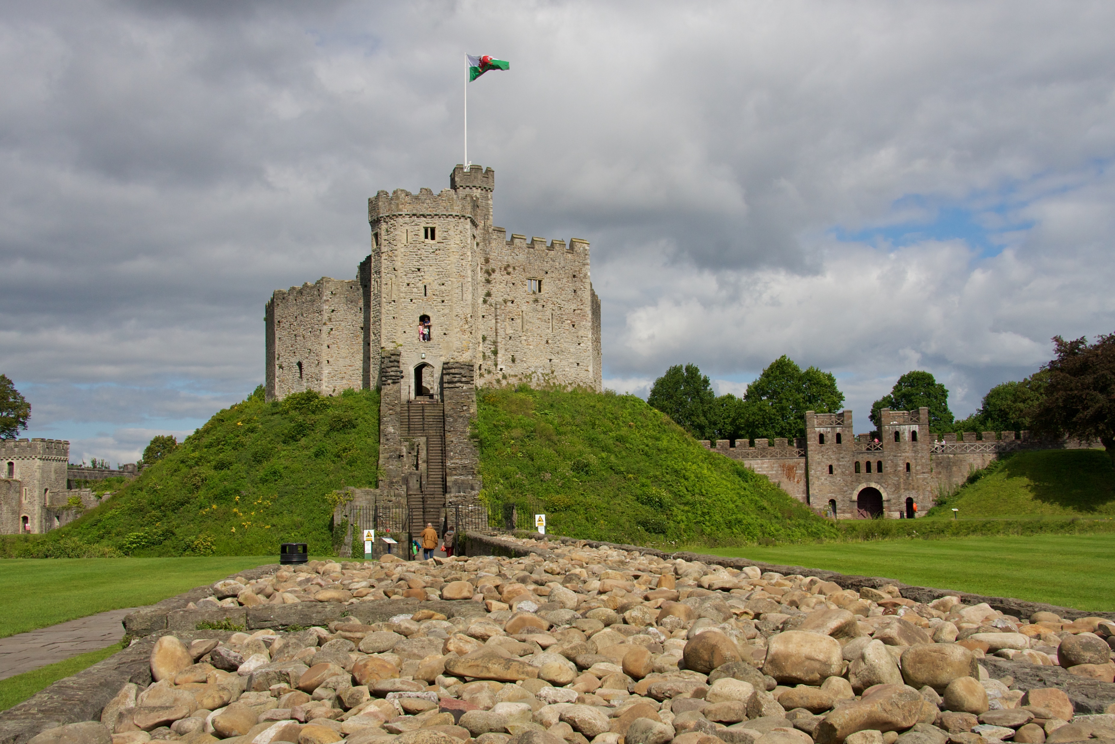 A stone medieval castle with a central keep stands atop a grassy mound, flying the Welsh flag. Ideal for students in Cathays seeking history, its rocky foreground and green lawns provide a scenic spot under cloudy skies.