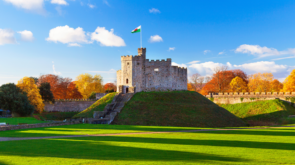 A stone castle with a central keep sits atop a grassy mound, surrounded by autumn trees under a blue sky. Ideal for students in Cathays seeking adventure, the site’s red dragon flag and sunlit lawns invite exploration and relaxation.