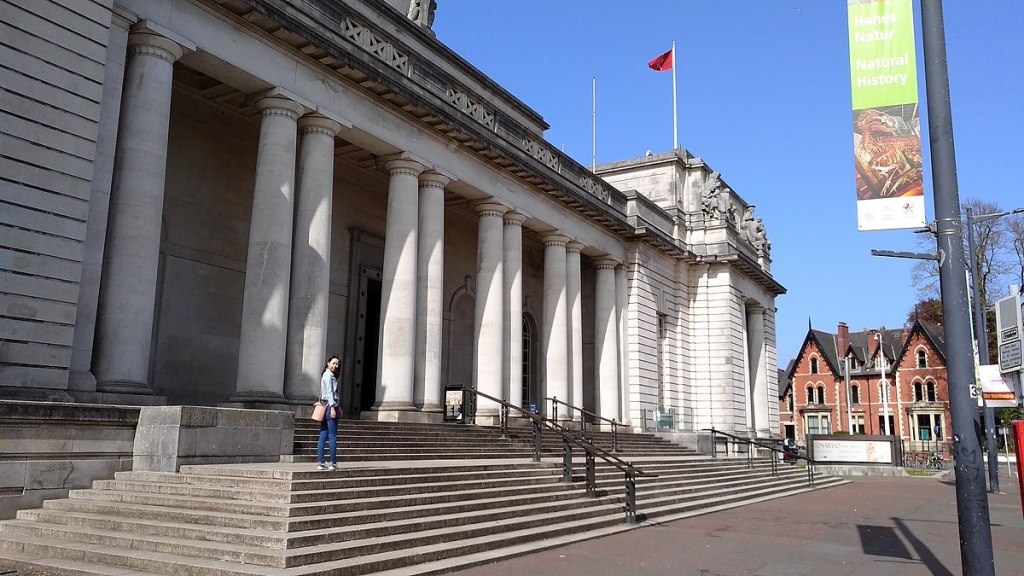 A person stands on the steps of a grand building with tall columns and a red flag on the roof—a must-see for students in Cathays looking for things to do in Cathays. A natural history banner and brick houses complete the scene under a clear blue sky.