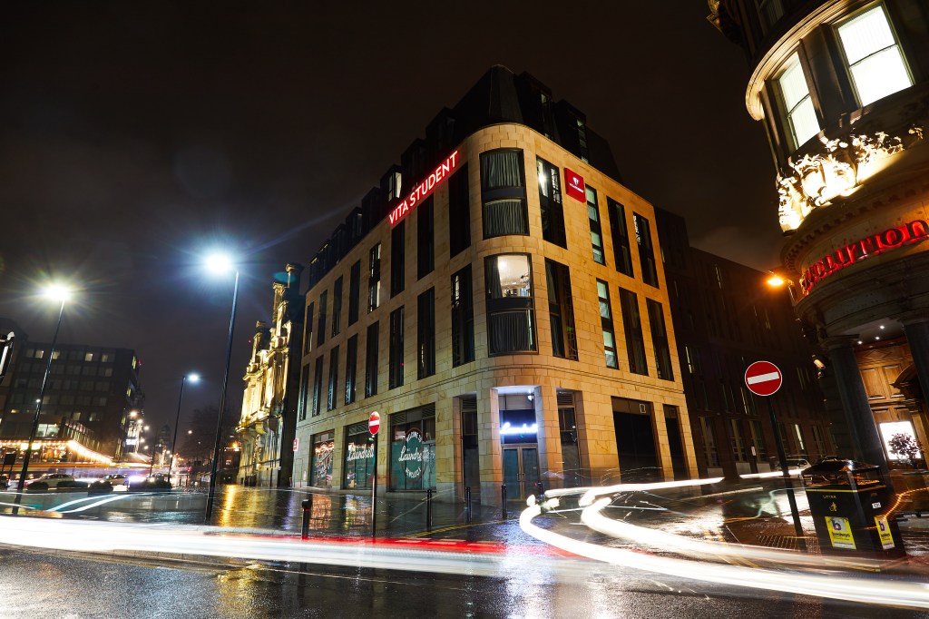 A modern, brightly lit corner building with "The Student" sign—an example of student accommodation Newcastle offers—seen at night with light trails from passing cars and wet streets reflecting city lights.