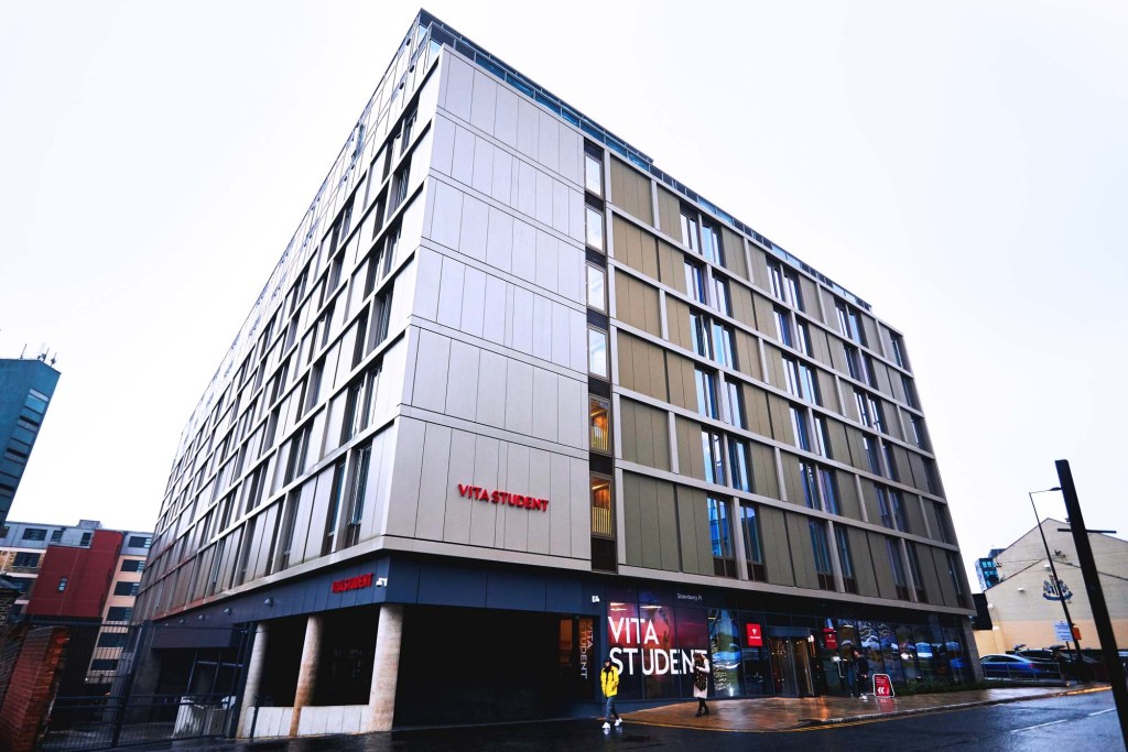 A modern, multi-story building with large windows and beige panels, labeled "Vita Student," showcases top student accommodation Newcastle offers. A person in a yellow jacket walks outside on a wet street by the glass doors and signs.