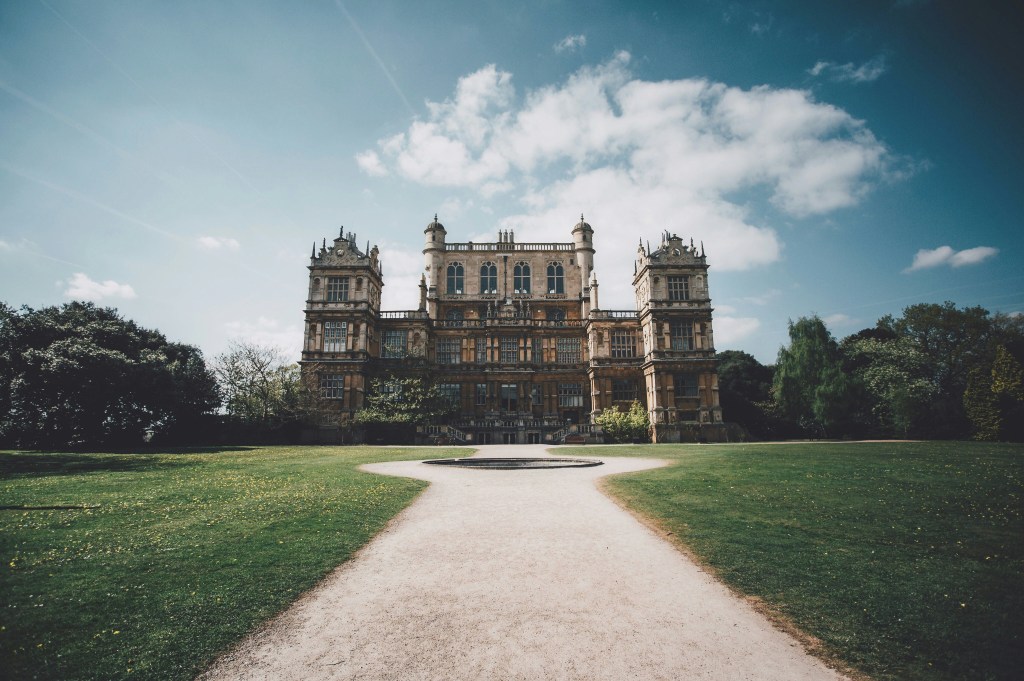 A grand, historic mansion with ornate architecture stands at the end of a wide gravel path, surrounded by green lawns and trees under a partly cloudy blue sky—a perfect spot for students in Nottingham to enjoy memorable days out.