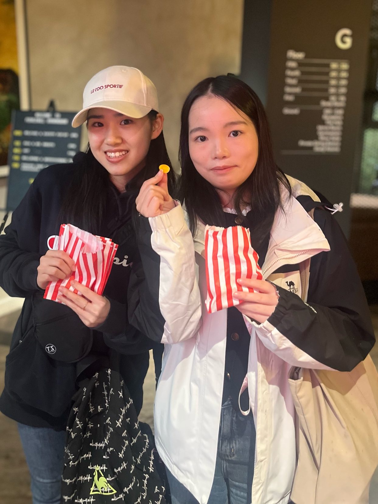 Two young women stand indoors, smiling and holding red-and-white striped bags. One woman holds a yellow candy. Both wear jackets; one has a white cap. The background features art and signage, hinting at fun Birmingham activities.