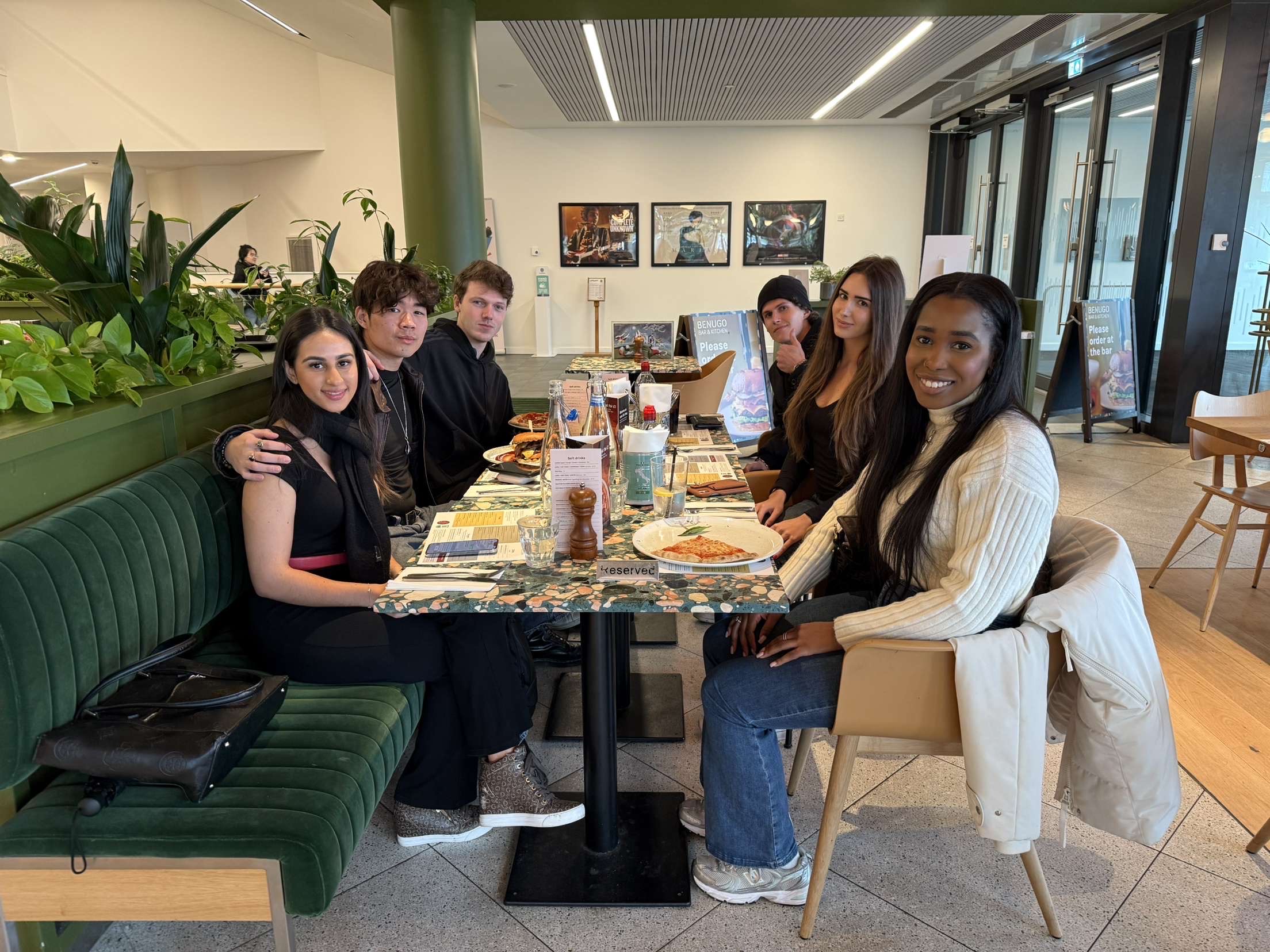 Six people, perhaps sharing student stories, sit around a table in a modern Vita Student café. Smiling at the camera, they enjoy plates of food and drinks in a bright setting with green plants and framed pictures on the wall.