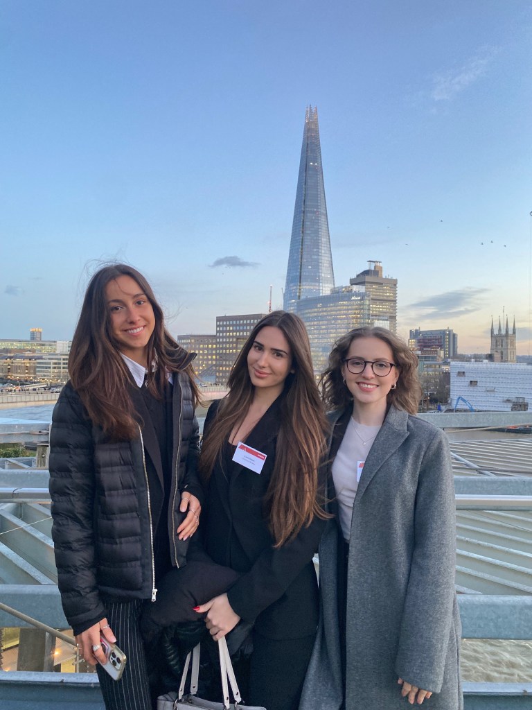 Three women stand smiling on a balcony with the Shard and London cityscape in the background. Wearing business attire and name badges, they share student experiences and stories of their unexpected route under a clear, early evening sky.