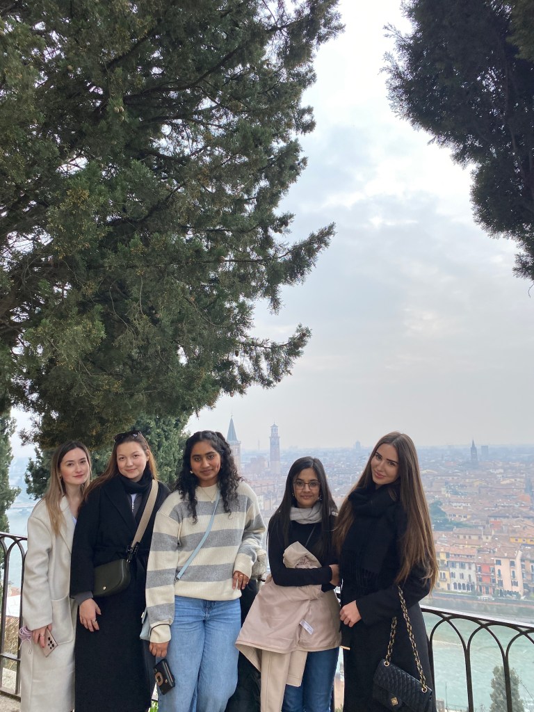 Five women stand together in front of a railing, surrounded by large evergreen trees and a scenic cityscape beneath a cloudy sky—a perfect moment capturing the spirit of student experiences and unexpected journeys.
