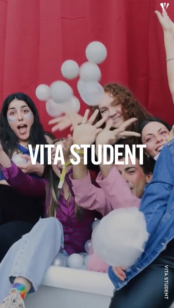 Five smiling young adults sit together in a tub filled with white balls, playfully throwing some up. A red curtain is behind them. Text across the image reads “VITA STUDENT.”.