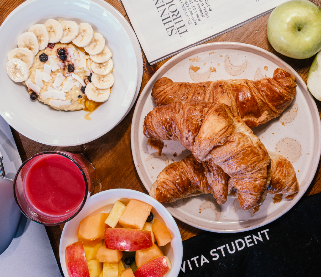 A breakfast spread in City Madrid with croissants, oatmeal topped with bananas and berries, a bowl of mixed fruit, red juice, green apples, and a newspaper on a wooden table.