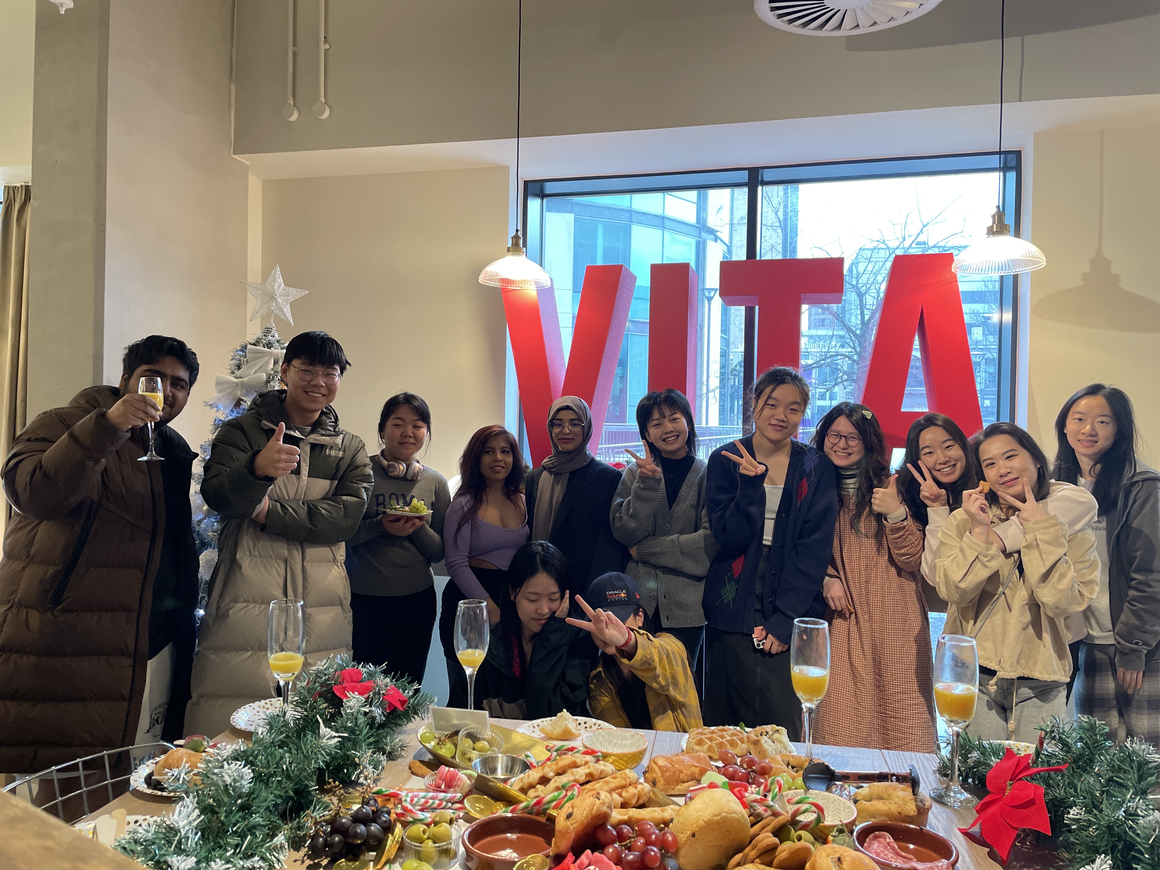 A group of twelve people stand and sit together, smiling in front of a holiday-themed table with food and drinks. Large red letters spelling "VITA" and a decorated tree highlight the Vita Student cultural celebration in the background.